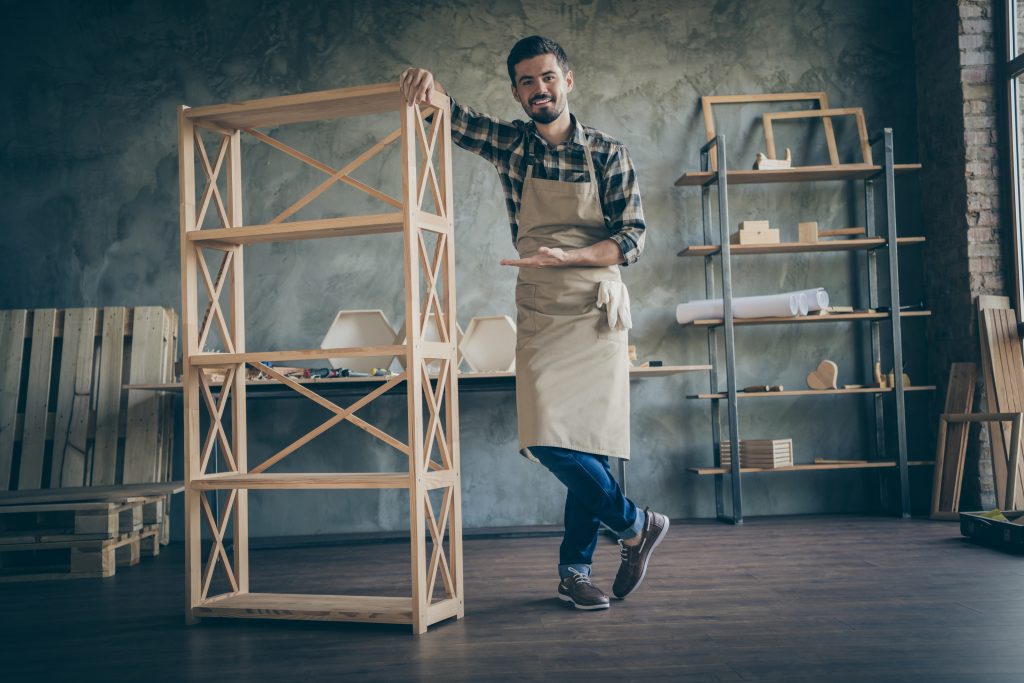 Full length photo of handsome cheerful guy showing just made bookshelf handmade, design wooden advertising own business industry woodwork shop indoors